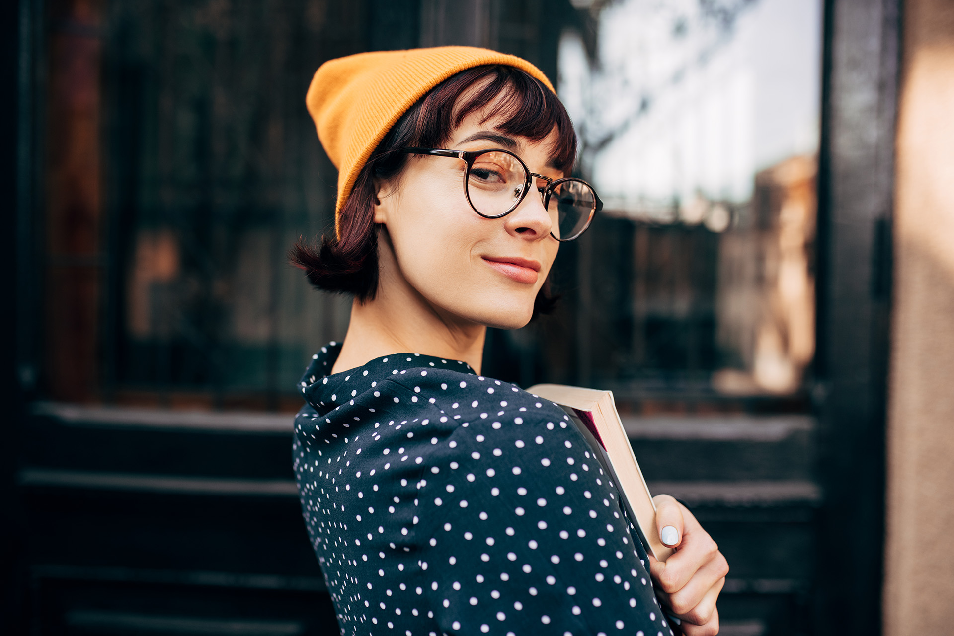 Eine Studentin mit einer gelben Wollmütze und Brille schaut in die Kamera und hält ihre UniBücher fest an die Brust gedrückt.