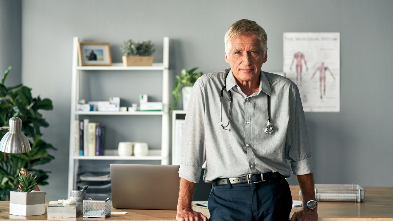 Älterer Mann mit grauem Hemd und Stethoskop lehnt an einem Schreibtisch in einem Büro mit Pflanzen und medizinischem Poster im Hintergrund
