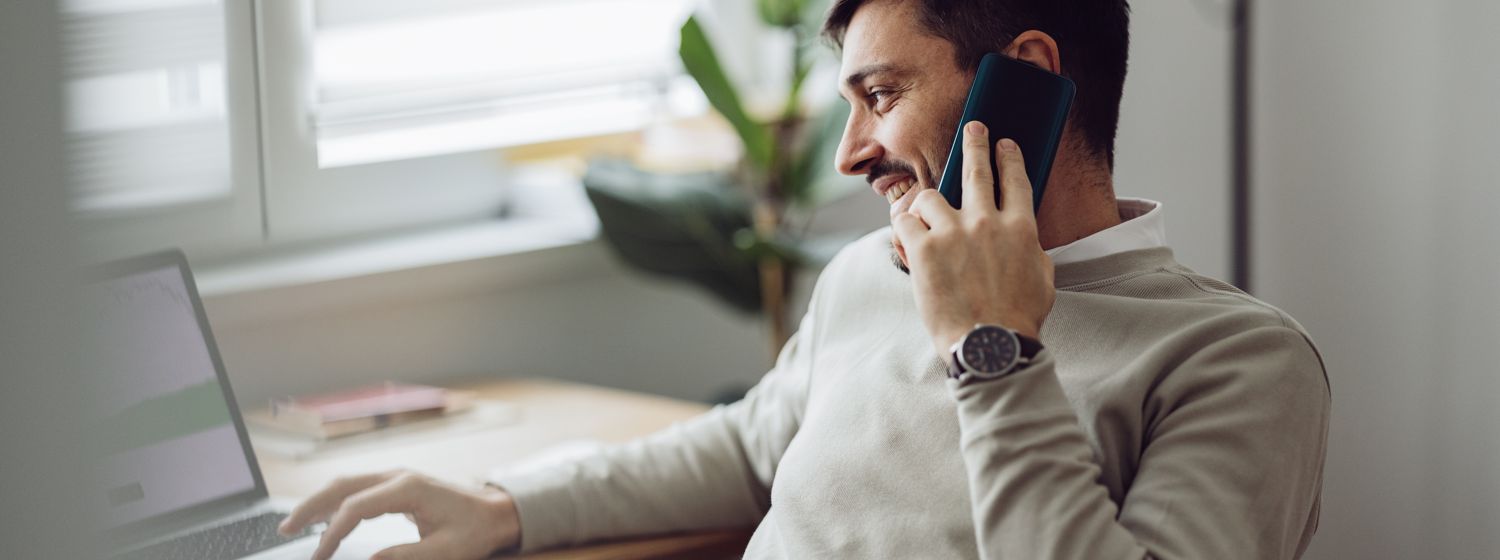 Mann am Laptop, telefonierend mit Smartphone am Ohr