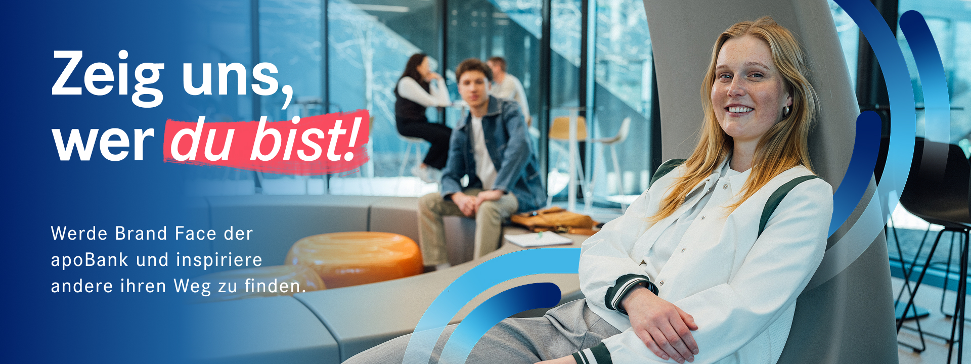 Eine Studentin mit einer weißen Collage-Jacke sitzt angelehnt in einem Raum auf dem Campus mit anderen Studentenschaut in die Kamera.